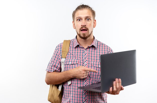 Удивленный молодой парень, студент, носящий рюкзак, держит и указывает на ноутбук, изолированный на белой стене