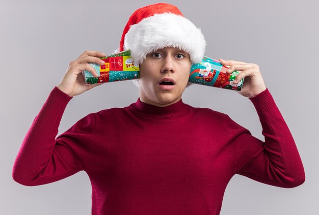 Surprised young guy wearing christmas hat holding christmas cups on ears isolated on white background