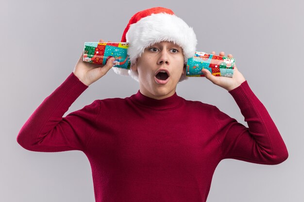 Surprised young guy wearing christmas hat holding christmas cups on ears isolated on white background