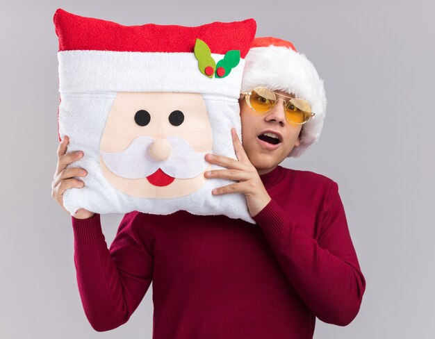 白い壁に分離されたクリスマスの枕を保持している眼鏡とクリスマスの帽子をかぶって驚いた若い男