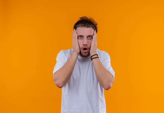 Surprised young guy wearing white t-shirt put his hands on cheeks on isolated orange background