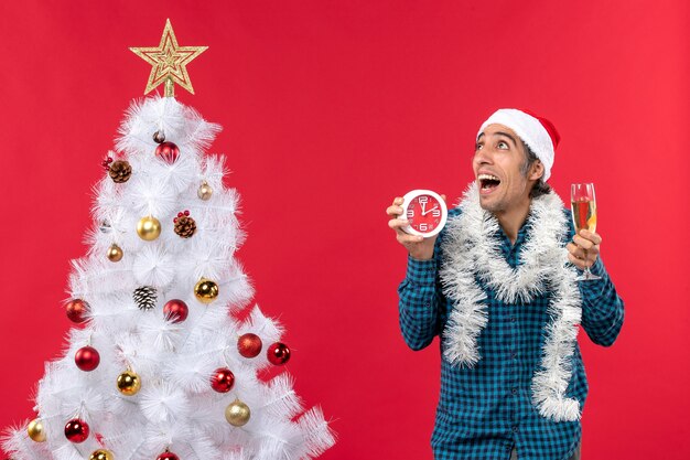 산타 클로스 모자와 크리스마스 트리 근처에 서있는 와인과 시계 한 잔을 들고 놀란 젊은 남자