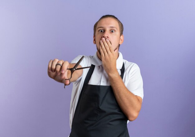 Free photo surprised young handsome barber wearing uniform putting hand on mouth and stretching out scissors towards front isolated on purple wall