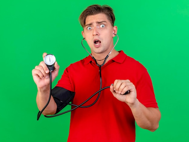 Surprised young handsome blonde ill man wearing stethoscope measuring pressure to himself showing sphygmomanometer looking at front isolated on green wall