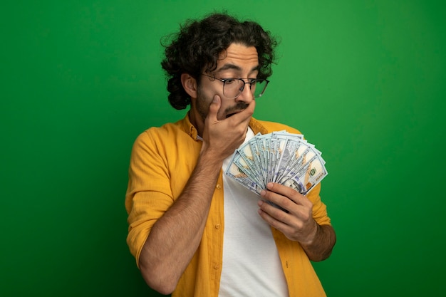 Бесплатное фото Удивленный молодой красивый кавказский мужчина в очках, держащий и смотрящий на деньги, держа руку за рот, изолированную на зеленой стене с копией пространства