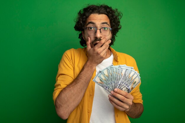 Surprised young handsome caucasian man wearing glasses holding money keeping hand on mouth  isolated on green wall with copy space