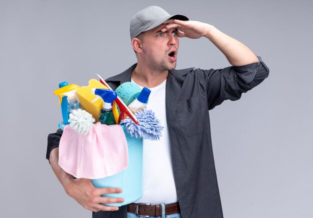 Surprised young handsome cleaning guy wearing t-shirt and cap holding bucket of cleaning tools looking at distance with hand isolated on white wall