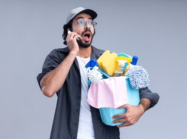 Surprised young handsome cleaning guy wearing t-shirt and cap holding bucket of cleaning tools and speaks on phone isolated on white wall