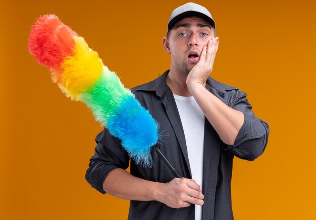 Surprised young handsome cleaning guy wearing t-shirt and cap holding pipidaster putting hand on cheek isolated on orange wall