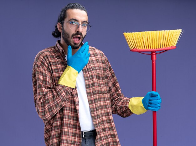 Surprised young handsome cleaning guy wearing t-shirt and gloves holding mop putting hand on chin isolated on blue wall