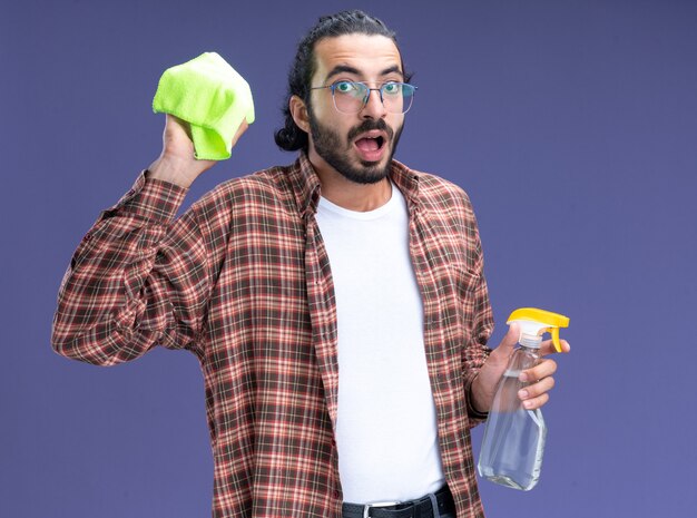 Surprised young handsome cleaning guy wearing t-shirt holding spray bottle with rag isolated on blue wall