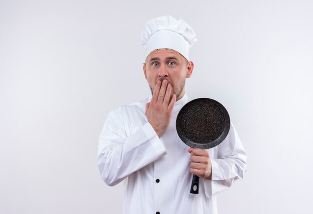 Surprised young handsome cook in chef uniform holding frying pan putting hand on mouth isolated on white space