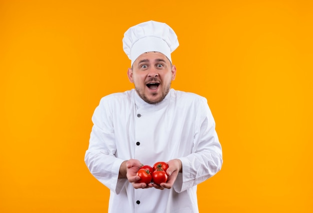 Surprised young handsome cook in chef uniform holding tomatoes isolated on orange space