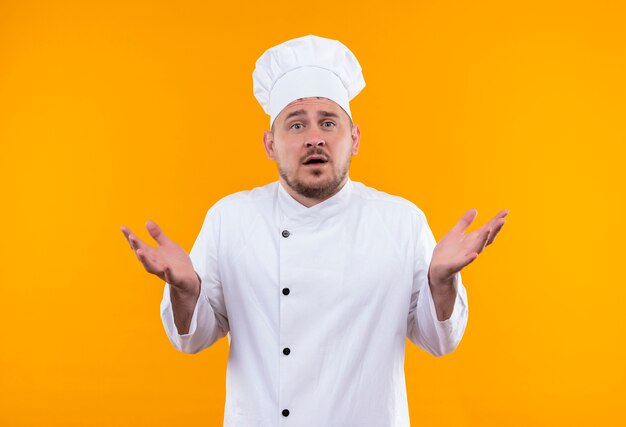Surprised young handsome cook in chef uniform showing empty hands on isolated orange space