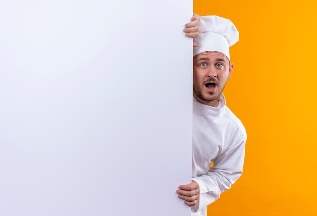 Удивленный молодой красивый повар в униформе шеф-повара стоит за белой стеной и держит ее изолированной на оранжевом пространстве