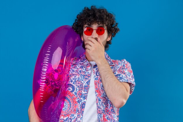 Surprised young handsome curly man wearing sunglasses holding swim ring with hand on mouth on isolated blue wall with copy space