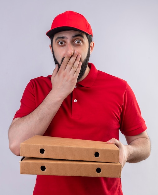 Удивленный молодой красивый курьер в красной форме и кепке держит коробки для пиццы и кладет руку на рот, изолированную на белой стене