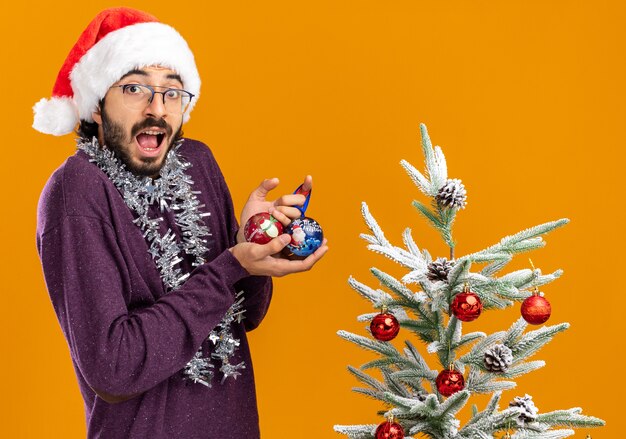 Surprised young handsome guy standing nearby christmas tree wearing christmas hat with garland on neck holding christmas balls isolated on orange background