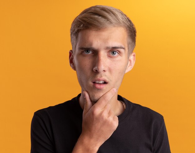 Surprised young handsome guy wearing black shirt grabbed chin isolated on yellow wall