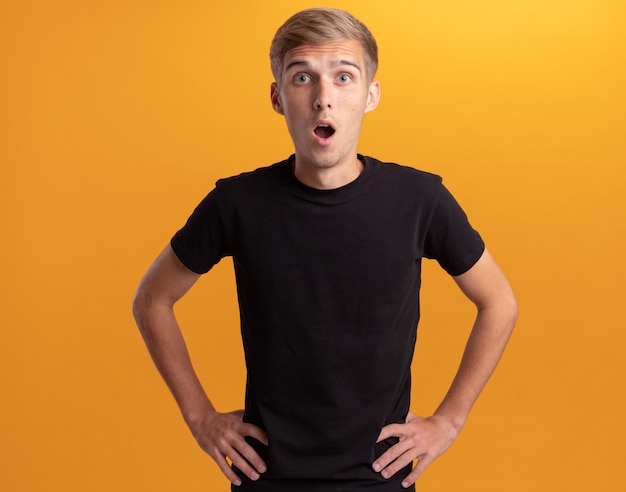 Surprised young handsome guy wearing black shirt putting hands on hip isolated on yellow wall