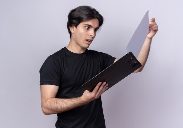 Surprised young handsome guy wearing black t-shirt flipping through clipboard isolated on white wall