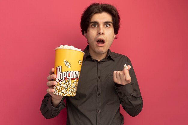 Surprised young handsome guy wearing black t-shirt holding bucket of popcorn and popcorn peace isolated on pink wall