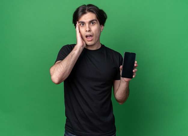 Surprised young handsome guy wearing black t-shirt holding phone and putting hand on cheek isolated on green wall