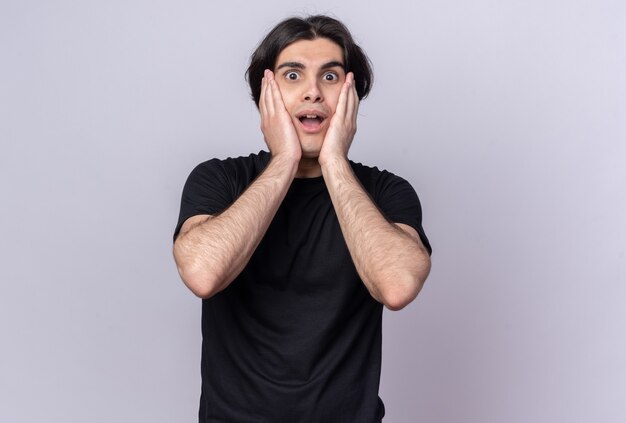 Surprised young handsome guy wearing black t-shirt putting hand on cheeks isolated on white wall