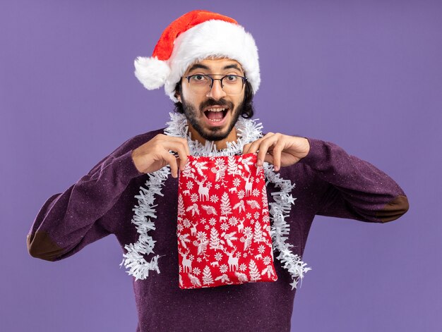 Surprised young handsome guy wearing christmas hat with garland on neck holding christmas bag isolated on blue background