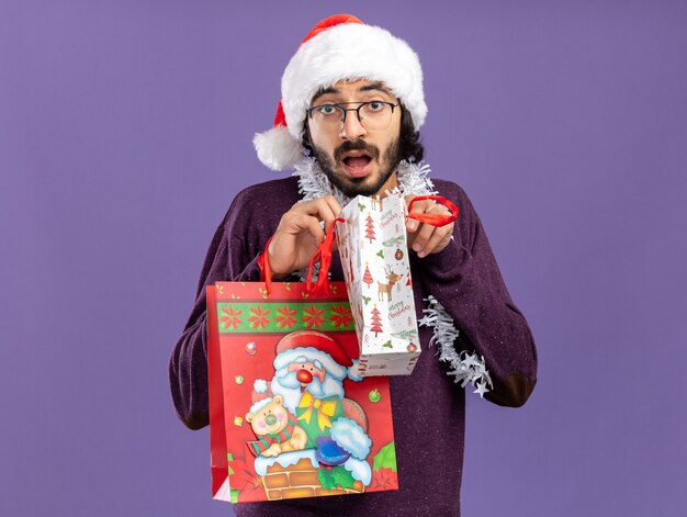 Surprised young handsome guy wearing christmas hat with garland on neck holding gift bags isolated on blue background