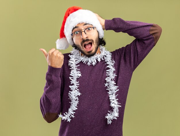 surprised young handsome guy wearing christmas hat with garland on neck points at side putting hand on head isolated on olive green wall with copy space