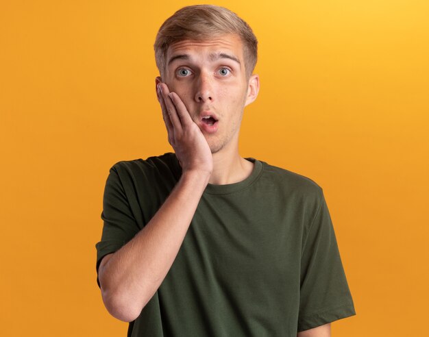 Surprised young handsome guy wearing green shirt putting hand on cheek isolated on yellow wall