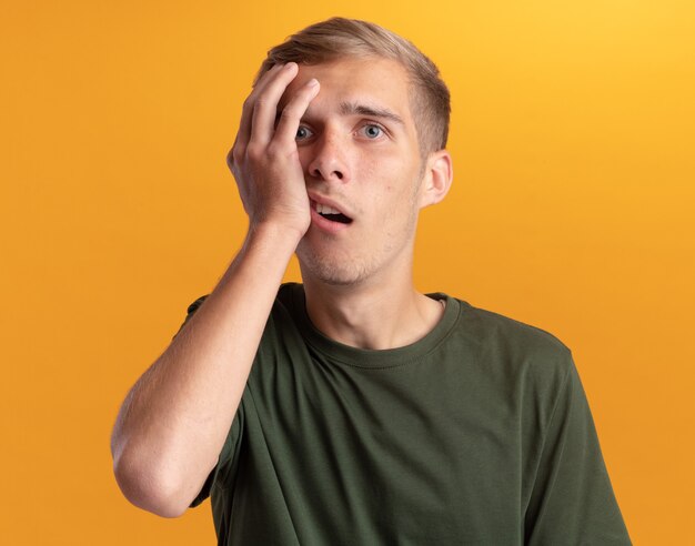 Surprised young handsome guy wearing green shirt putting hand on cheek isolated on yellow wall