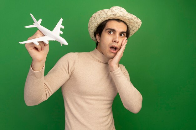 Surprised young handsome guy wearing hat and raising toy airplane putting hand on cheek isolated on green wall