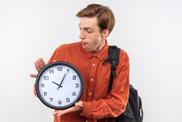 Sorpreso giovane bel ragazzo che indossa una camicia rossa con lo zaino che tiene e guarda l'orologio da parete isolato sul muro bianco