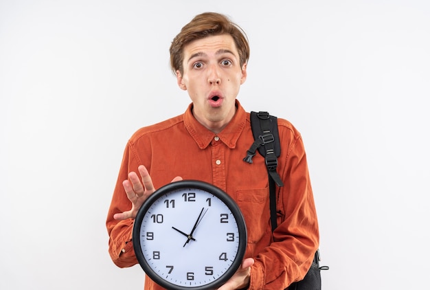 Surprised young handsome guy wearing red shirt with backpack holding wall clock isolated on white wall