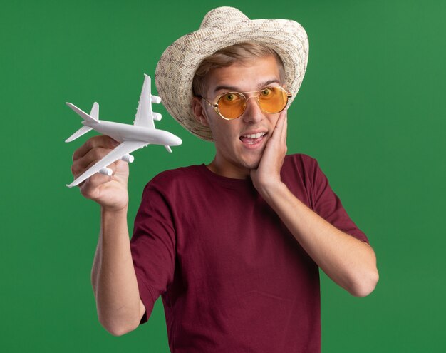 Surprised young handsome guy wearing red shirt with glasses and hat holding toy airplane putting hand on cheek isolated on green wall