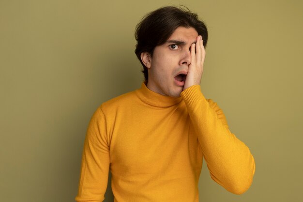 Surprised young handsome guy wearing yellow turtleneck sweater covered eye with hand isolated on olive green wall