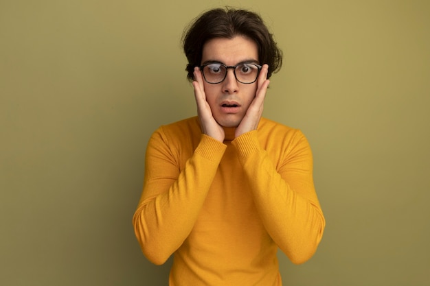 노란색 터틀넥 스웨터와 안경을 착용하고 올리브 녹색 벽에 고립 된 뺨에 손을 댔을 놀란 젊은 잘 생긴 남자