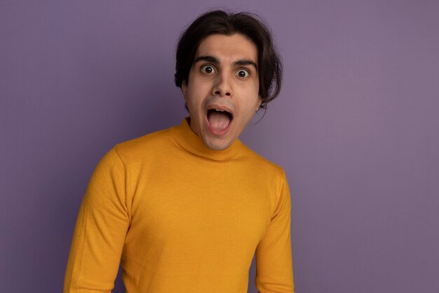 コピースペースと紫色の壁に分離された黄色のタートルネックのセーターを着て驚いた若いハンサムな男
