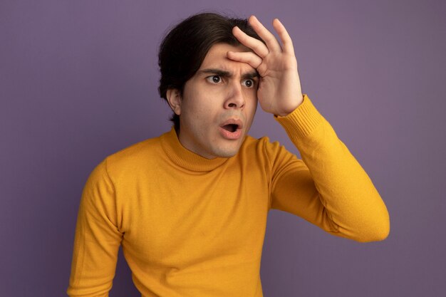 紫色の壁に隔離された手で距離を見て黄色のタートルネックのセーターを着て驚いた若いハンサムな男
