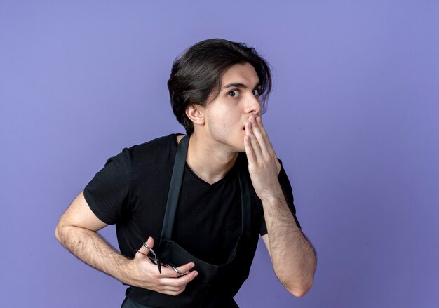 Surprised young handsome male barber in uniform holding scissors and covered mouth with hand isolated on blue