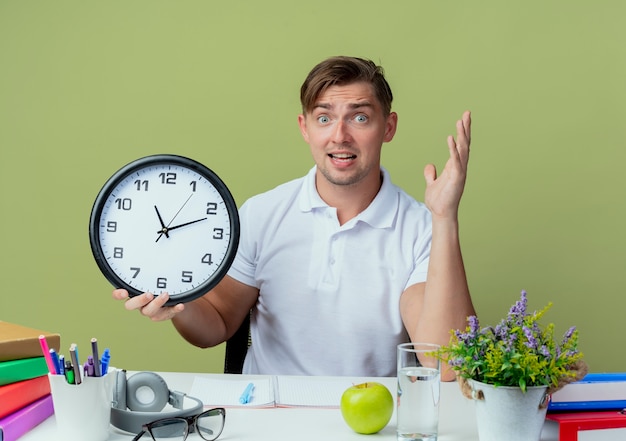 Giovane studente maschio bello sorpreso che si siede allo scrittorio con gli strumenti della scuola che tengono l'orologio di parete e la mano diffusa isolata su verde oliva