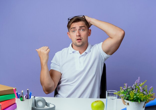 Surprised young handsome male student sitting at desk with school tools wearing glasses on head and putting hand on head points at side isolated on blue with copy space