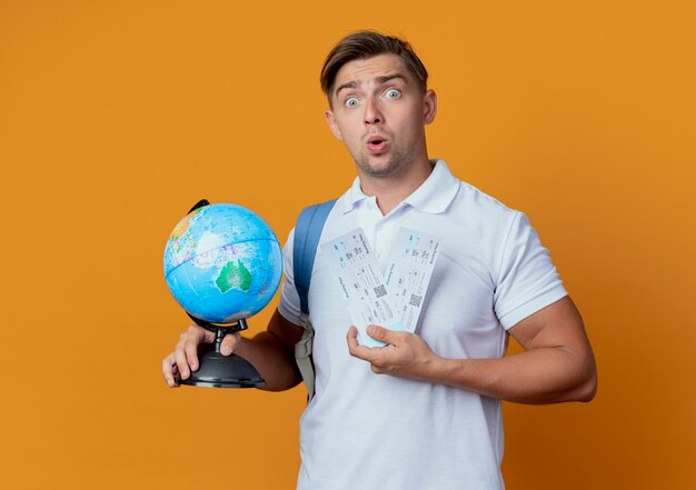 Surprised young handsome male student wearing back bag holding tickets and globe isolated on orange
