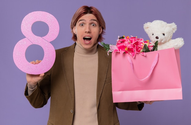 ピンクの8番と花とテディベアのギフトバッグを持って驚いた若いハンサムな男