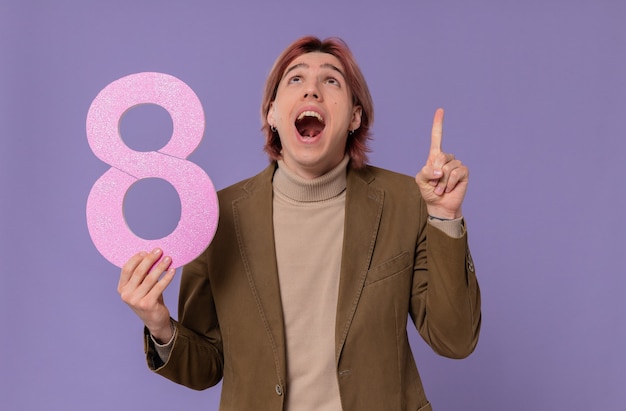 Surprised young handsome man holding pink number eight looking and pointing up 