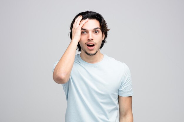 Surprised young handsome man looking at camera kepping hand on head isolated on white background