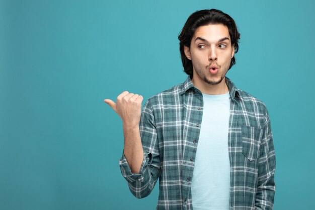 surprised young handsome man looking at camera pointing to side isolated on blue background with copy space