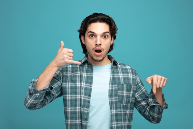 surprised young handsome man looking at camera showing call gesture pointing down isolated on blue background